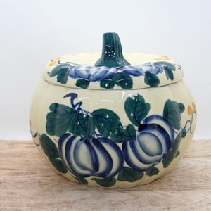 Peut inclure: Un pot en céramique crème en forme de citrouille avec un couvercle, orné de citrouilles peintes à la main en bleu et blanc, de feuilles vert foncé et de petits accents jaunes. Le couvercle présente une tige verte et une texture côtelée.