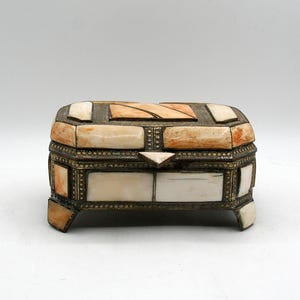 May include: An ornate, rectangular jewelry box with a hinged lid. The box features a cream and orange mosaic design, accented with brass-colored metal trim and small feet. The box is likely used for storing small items.