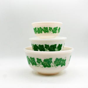 May include: A set of three stacked off-white glass bowls of varying sizes, each featuring a repeating pattern of vibrant green ivy leaves and thin curling vines encircling the exterior rim of the bowls against a smooth, opaque glass surface.