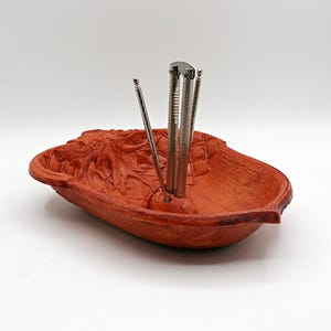 May include: A reddish-brown wooden bowl carved in the shape of an acorn with a textured cap pattern. Three silver-colored metal nut picks are inserted into a small hole in the center of the bowl, standing upright against the interior surface.