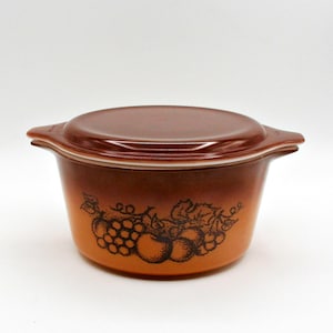 Puede incluir: Una fuente para horno vintage marrón y naranja con tapa a juego. La fuente presenta un diseño de frutas con uvas y manzanas. La fuente para horno tiene asas y es de cerámica.