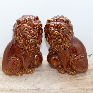 May include: Two brown ceramic lion statues. The lions are seated, with detailed manes and expressive faces. The statues have a glossy finish and are displayed on a light-colored wooden surface. These decorative figurines add a touch of elegance.