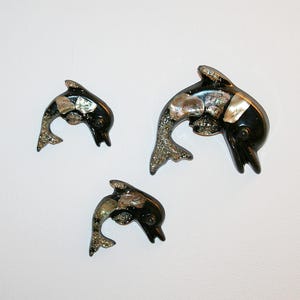 Puede incluir: Tres figuras decorativas de delfines. Cada delfín es negro con detalles iridiscentes y una cola plateada brillante. Los delfines varían en tamaño y están dispuestos sobre un fondo blanco. Ideales para decoración de pared o como acentos decorativos.