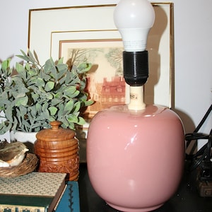 Op de afbeelding: Een tafellamp met een gladde, ronde, oudroze keramische voet en een witte gloeilamp. De lamp heeft een zwarte fitting en een crèmekleurige hals. De voet is glanzend en bolvormig en staat rechtop zonder een lampenkap.