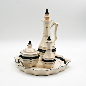 Puede incluir: Un juego de servir de cerámica color crema con rayas geométricas negras. El juego incluye una jarra alta, un tarro pequeño con tapa y una botella pequeña, todo sobre una bandeja festoneada. La jarra tiene un asa y la letra "V" en un lado.