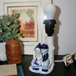 May include: A ceramic table lamp featuring a white and blue figurine of two children sitting on a bench against a tree trunk base. The base is marked with the word HOLLAND. The lamp includes a black socket and a white light bulb on top.