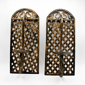 Peut inclure: Une paire de bougeoirs muraux dotés d'un cadre rectangulaire avec un sommet arqué et un motif en treillis tressé. La structure en métal présente une finition noire avec des reflets dorés vieillis et une plateforme circulaire pour bougies.