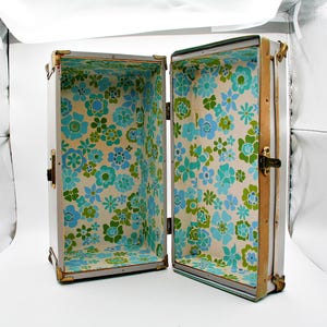 May include: An open vintage trunk with a white exterior and brass hardware. The interior is lined with a floral pattern in shades of blue and green on a cream background. The trunk is rectangular and has a hinged lid.