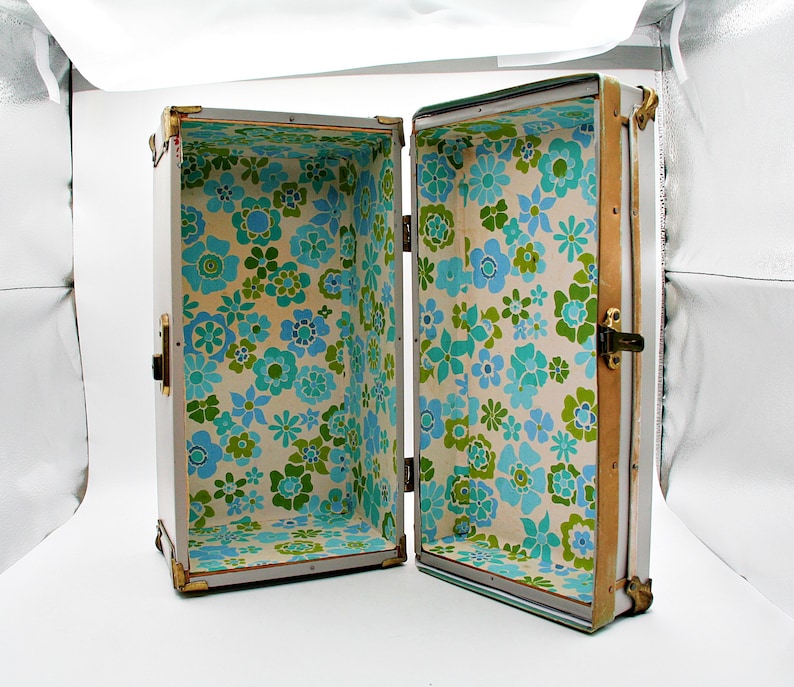 May include: An open vintage trunk with a white exterior and brass hardware. The interior is lined with a floral pattern in shades of blue and green on a cream background. The trunk is rectangular and has a hinged lid.