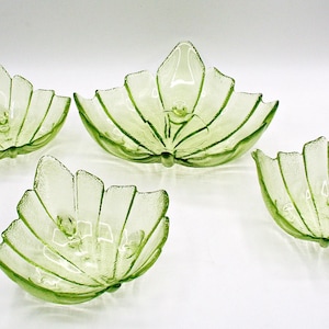 May include: A set of four light green glass bowls shaped like leaves with textured edges and ribbed details. The collection includes one large central bowl and three smaller bowls arranged around it, all featuring a translucent finish.