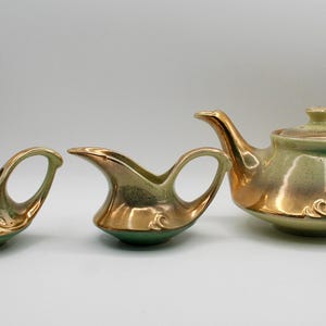 Puede incluir: Un juego de té vintage con tetera, crema y azucarero. El juego presenta una combinación de colores dorados y verdes con un acabado brillante. La tetera tiene tapa y asa curva. El azucarero tiene dos asas.