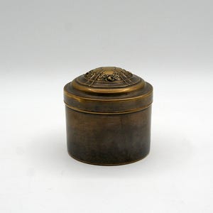 Peut inclure: Un récipient cylindrique en laiton avec une finition métallique sombre et lisse, doté d'un couvercle amovible. Le dessus du couvercle présente un motif en relief circulaire avec des détails floraux et des lignes géométriques gravées.