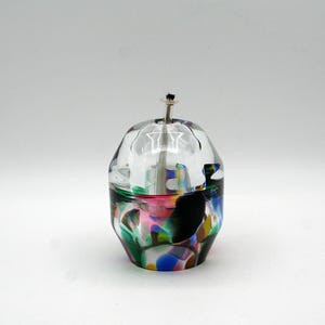 May include: A decorative glass oil lamp with a clear top and a colorful base. The base features a mix of colors including green, pink, blue, and black. A silver wick holder extends from the top.
