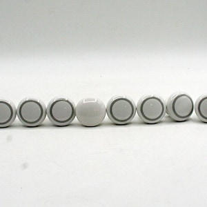 May include: A set of twelve round cabinet knobs arranged in a horizontal line. Eleven of the knobs are white with a thin grey circular ring detail, while one knob in the centre is solid white without any additional markings or patterns.
