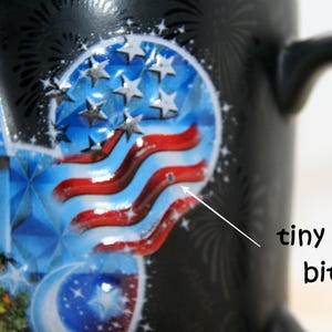 May include: Black ceramic mug with a patriotic design featuring a stylized American flag, stars, and a blue castle. The mug has a black handle and a small imperfection. The mug is decorated with fireworks.