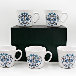 Puede incluir: Un juego de cinco tazas de cerámica blanca, cada una con un patrón floral simétrico en azul y marrón con aves estilizadas. Las tazas tienen un asa redondeada y una forma ligeramente cónica, mostrando un motivo decorativo consistente.