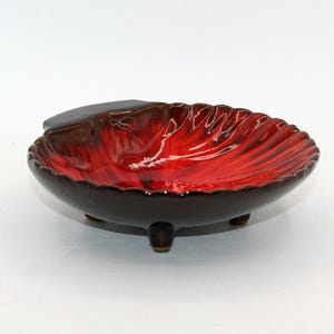 Może przedstawiać: Ceramiczna miseczka w kształcie muszli z błyszczącym wykończeniem. Wnętrze ma żywy czerwony gradient, który przechodzi w głęboki ciemny brąz na krawędziach. Zewnętrzna strona jest ciemnobrązowa, a naczynie stoi na trzech małych nóżkach.