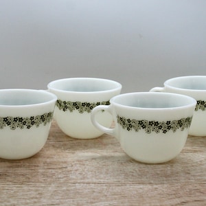 Pode incluir: Quatro xícaras de chá brancas com um desenho floral em preto e verde. As xícaras têm formato arredondado com alça e uma faixa decorativa de flores na parte superior. As xícaras estão dispostas sobre uma superfície de madeira.
