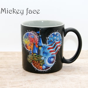 May include: Black ceramic mug with a Mickey Mouse face design. The design features a castle, fireworks, an American flag, and a crescent moon. The mug has a black handle and a gray interior. The text "Mickey face" is above the mug.