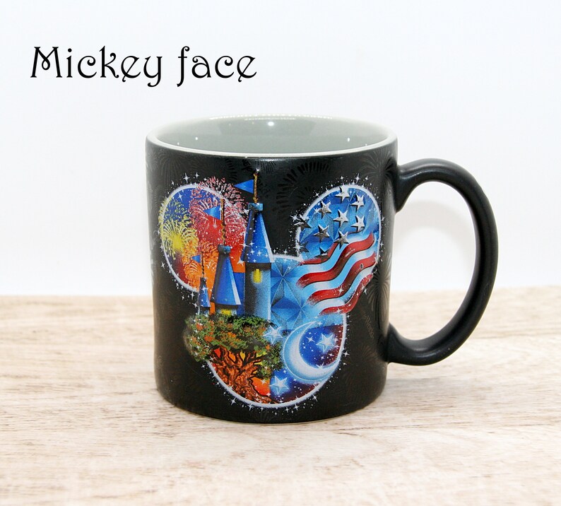 May include: Black ceramic mug with a Mickey Mouse face design. The design features a castle, fireworks, an American flag, and a crescent moon. The mug has a black handle and a gray interior. The text "Mickey face" is above the mug.