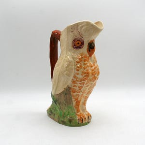 May include: A ceramic pitcher shaped like an owl with a cream-colored body, orange textured chest feathers, and a dark brown handle. The owl has large orange eyes, a dark beak, and stands on a green base. The spout is formed by the owl's head.