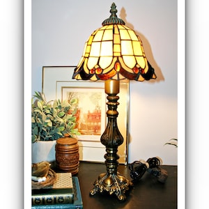 May include: A table lamp with a stained glass shade featuring cream-colored rectangular panels and amber-toned accents. The lamp has a dark, ornate metal base with intricate detailing and a finial at the top. The light emits a warm, soft glow.