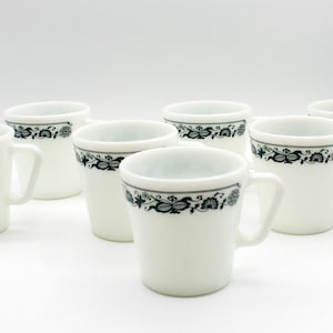 Pode incluir: Conjunto de oito canecas de cerâmica branca com um design floral preto na borda. Cada caneca tem uma alça e uma forma ligeiramente alargada. As canecas estão dispostas sobre uma superfície branca.