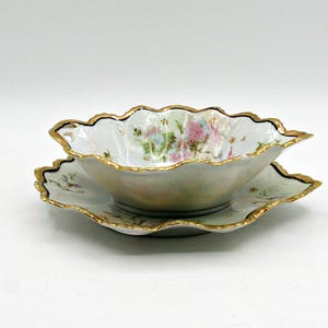 May include: A white porcelain bowl and matching saucer set featuring a delicate floral pattern in pink and blue hues. Both pieces have a scalloped edge finished with a thin gold trim and a dark blue accent line along the scalloped rim.