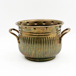 May include: A round brass planter with a weathered, tarnished patina. The vessel features vertical ribbed detailing along the body, two curved side handles, and a decorative rim with a series of small, evenly spaced circular indentations.