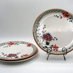 May include: Set of three white ceramic plates with a floral design. The plates feature a brown rim, a green and brown border, and red and purple flowers. One plate is on a black metal stand.