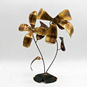 Puede incluir: Una escultura de flores de metal hecha a mano con dos grandes flores estilizadas y varias hojas. Las flores y las hojas tienen un tono bronce dorado cálido, mientras que los tallos y la base son de color marrón oscuro. Es una pieza decorativa.
