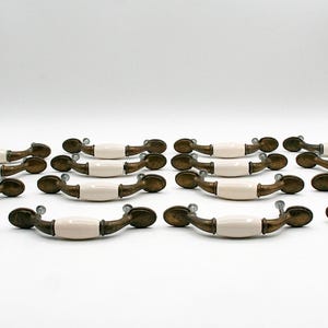 May include: A collection of antique cabinet pulls. Each pull features a cream-colored ceramic center, brass-toned hardware, and a curved design. The pulls are arranged in rows on a white surface, showcasing their vintage aesthetic.