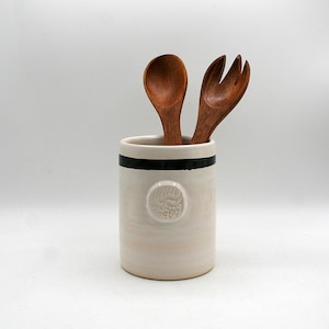 May include: A cream ceramic utensil holder with a black stripe and embossed design, containing a wooden spoon and fork. The wooden utensils are a warm brown, contrasting with the off-white holder. The cylindrical holder sits on a white surface. The holder is approximately 15 cm tall.