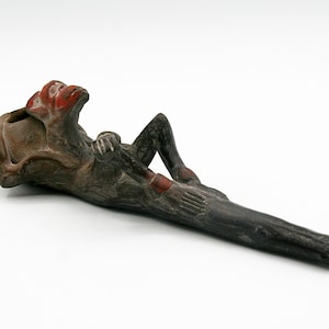 Puede incluir: Una pipa de madera tallada que representa una figura reclinada con la cara y las manos rojas. La pipa es principalmente de color marrón oscuro con detalles en rojo, la figura está tumbada en el tallo.