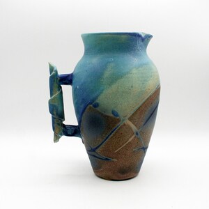 May include: A ceramic pitcher featuring a textured surface with a gradient of blue, teal, and earthy brown tones. The vessel has a wide mouth, a tapered base, and a distinctively sculpted, angular handle with layered, folded clay elements.