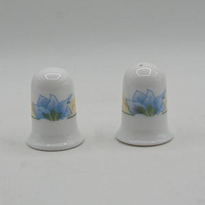 May include: Two white ceramic thimbles featuring a painted blue flower design with green leaves and yellow accents. Each thimble has a smooth, rounded top and a flared base, displaying a consistent floral pattern around the centre of the body.