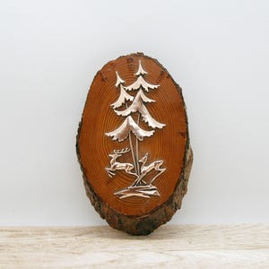 May include: A vertical oval-shaped wood slice plaque featuring a polished silver-toned metal inlay of two pine trees and two deer standing at the base. The wood displays a natural brown grain pattern with a rough, textured bark edge on the sides.