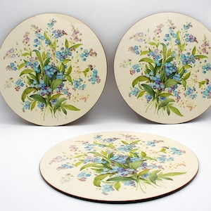 Peut inclure: Trois dessous de plat ronds avec un motif floral. Les dessous de plat ont un fond crème avec un bouquet de fleurs bleues, violettes et vertes. Les bords des dessous de plat sont de couleur marron foncé.