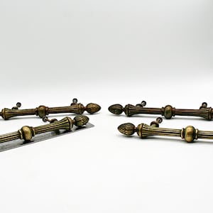 May include: Six antique bronze cabinet pulls with a decorative design. Each pull features a central cylindrical body with vertical grooves, flanked by decorative finials. A ruler is placed near the pulls for scale.