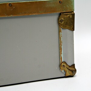 May include: Close-up of a gray storage box with brass-colored metal trim and corner protectors. The metal has a distressed, aged appearance. The box is set against a white background.