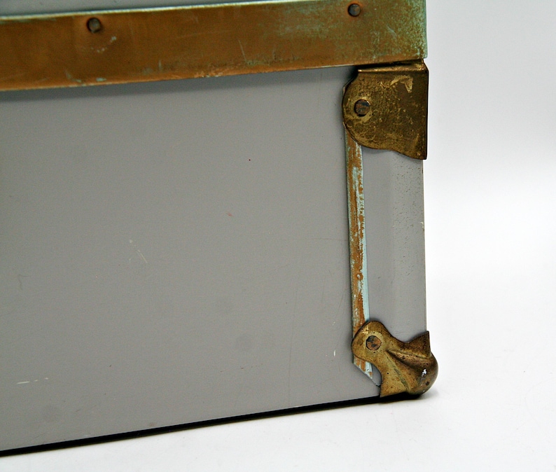 May include: Close-up of a gray storage box with brass-colored metal trim and corner protectors. The metal has a distressed, aged appearance. The box is set against a white background.