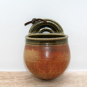 May include: A wall-mounted ceramic salt pig with a rounded body featuring a gradient of earthy orange and brown tones. The top rim and arched hanging tab are finished in a dark olive green glaze. A brown leather cord is tied through the hanging tab.