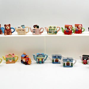 Può includere: Una collezione di teiere decorative con vari disegni, tra cui motivi di orsi, case e treni. Le teiere sono dai colori vivaci e realizzate in ceramica. Alcune teiere hanno un'altezza di circa 7,6 cm.