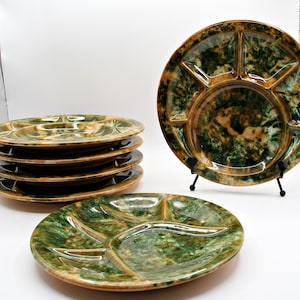 May include: A collection of six divided serving plates with a speckled green and brown glaze. One plate is displayed on a black stand. The plates have a central round section and four pie-shaped compartments.