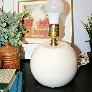 Puede incluir: Una base de lámpara esférica de cerámica color crema con un casquillo de metal tono latón y una bombilla LED blanca. La lámpara presenta un acabado liso y brillante con una forma redondeada, colocada en posición vertical con un cable blanco.