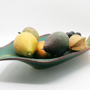 May include: A decorative leaf-shaped ceramic bowl in shades of green and red, filled with various faux fruits. The fruits include a yellow lemon, a green avocado, and a star fruit. The bowl is on a white background.