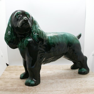 Può includere: Statuetta di cane in ceramica con una glassa verde scuro lucida. Il cane è raffigurato in posizione eretta, con orecchie lunghe e flosce e una coda corta. La statuetta è esposta su una superficie di legno chiaro.