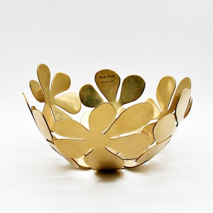 May include: A gold-toned metal bowl constructed from multiple overlapping flower-shaped petals. The interior surface features the engraved text "Monika Mulder" and "Design House Stockholm". The bowl has a rounded, open form with a smooth finish.