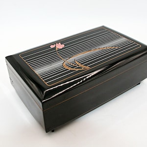 May include: A rectangular black box with a glossy finish featuring a pink flower illustration and thin gold horizontal lines on the lid. The box has a hinged lid and small feet at the base, presenting a smooth, dark surface with decorative gold accents.