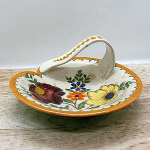 May include: A ceramic dish with a curved handle, featuring a cream-colored base with a hand-painted floral design in red, yellow, orange, blue, and green. The rim is painted in a solid orange color, and the handle has a small yellow leaf pattern.